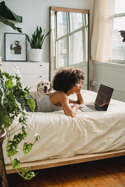 Woman laying on a bed with her dog next to her. She's viewing a laptop screen that's in front of her.