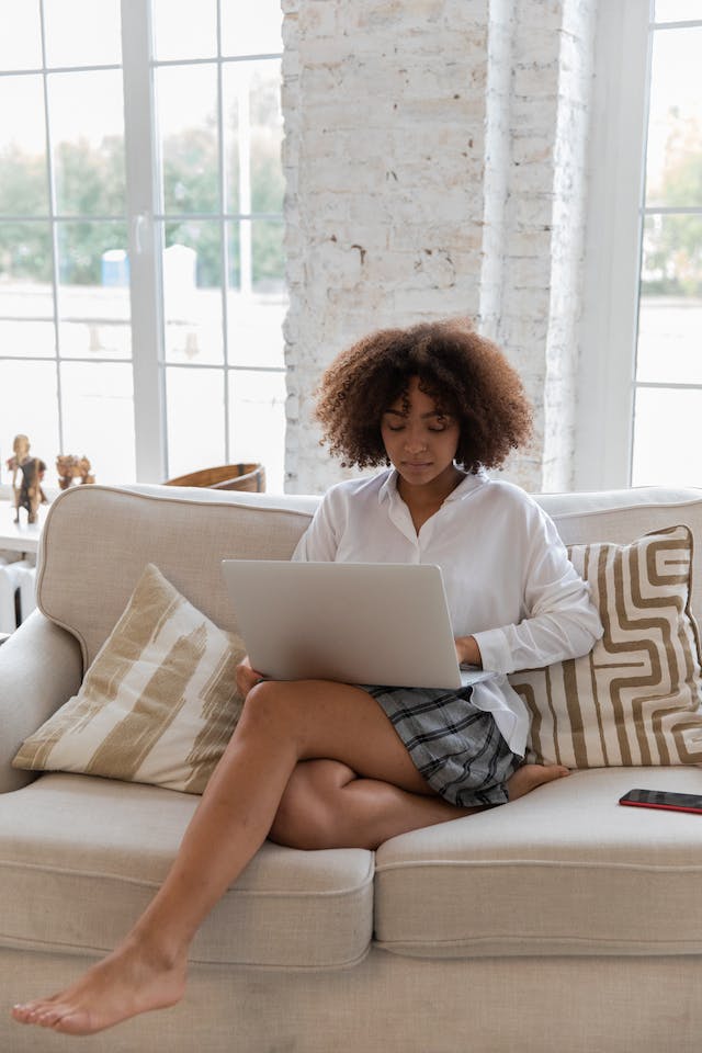 Woman with curly hair sitting on a couch viewing the laptop that's on her lap.